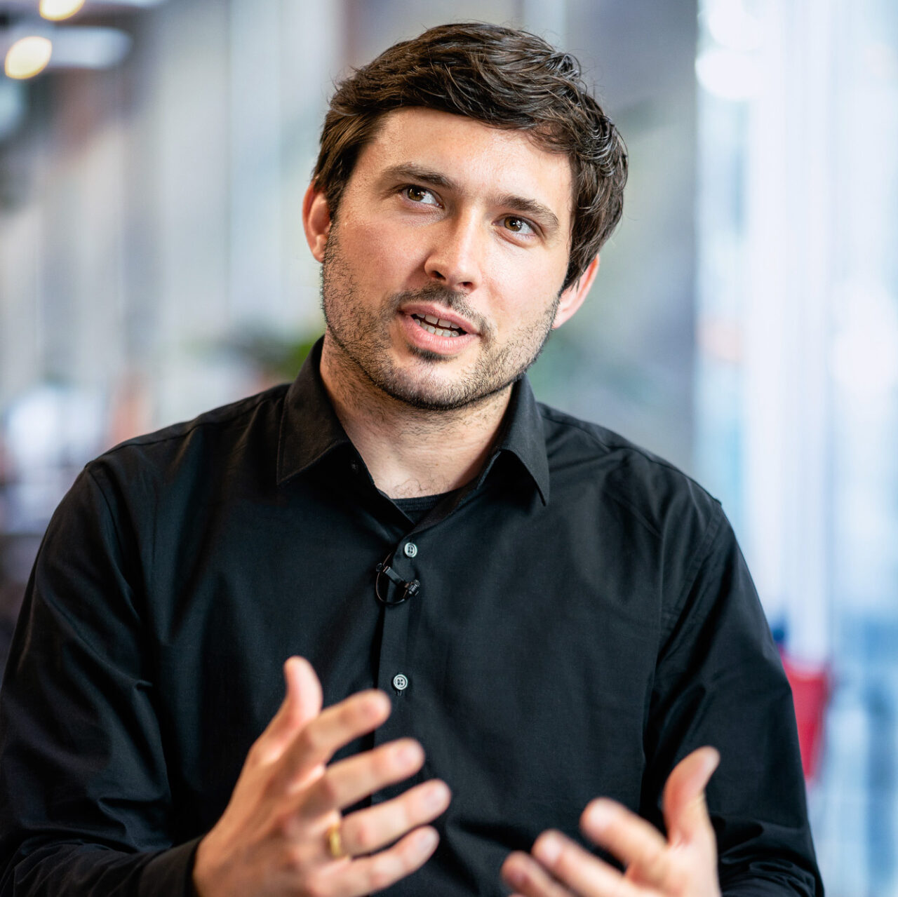 Homme aux cheveux bruns, chemise noir, qui à l'air d'expliquer quelque chose